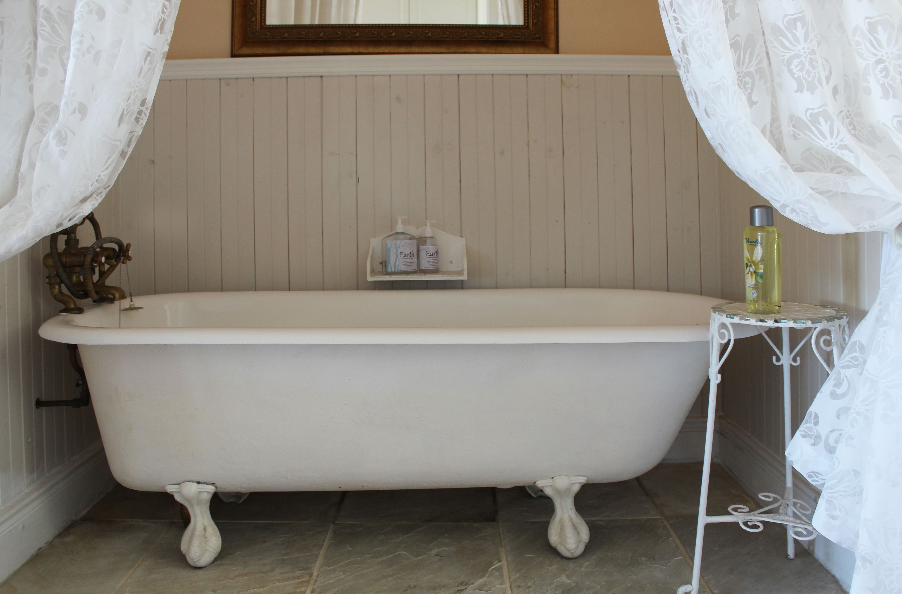 A white clawfoot bathtub with a wooden panel wall, a small shelf with toiletries, and a metal side table with a bottle.
