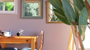 A room with a wooden table and chairs, a vase with green leaves, and a window with a view of trees. - Bluebottle Guest House (Cape Town)