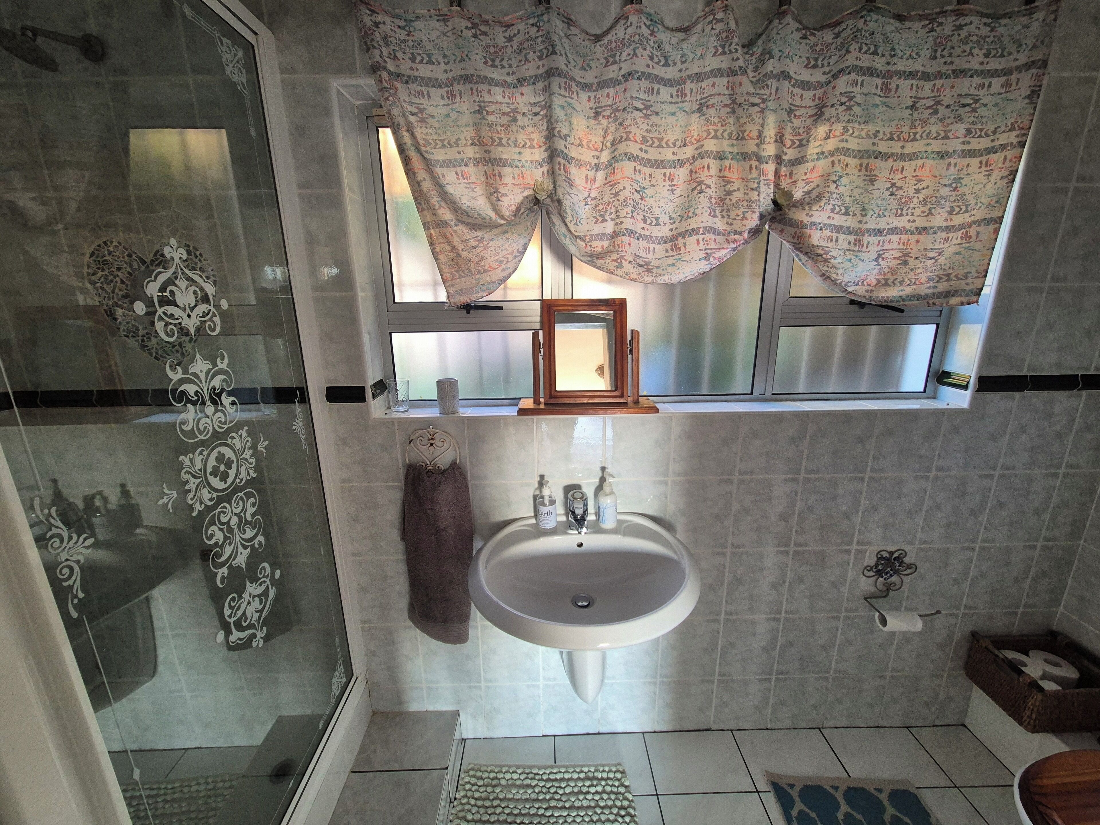 A bathroom with a glass shower enclosure, a sink with a mirror above, patterned curtains, and a small window.