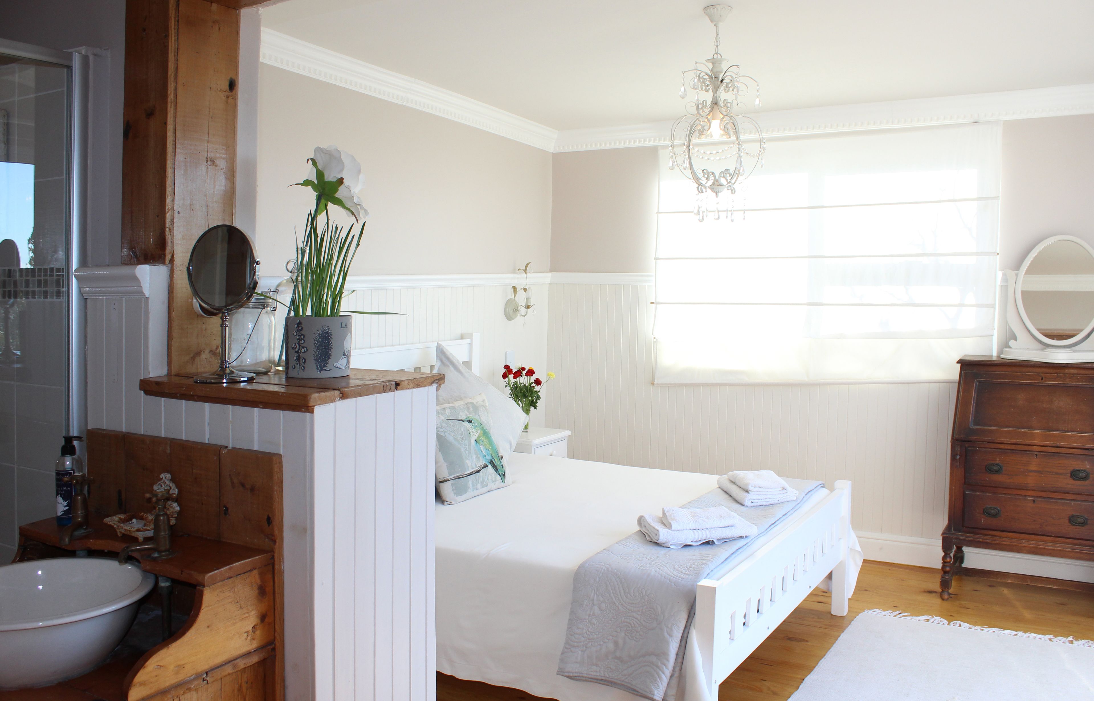 A bedroom with a wooden dresser, a bedside table with a mirror, a white bed with a white blanket, and a window with sheer curtains.
