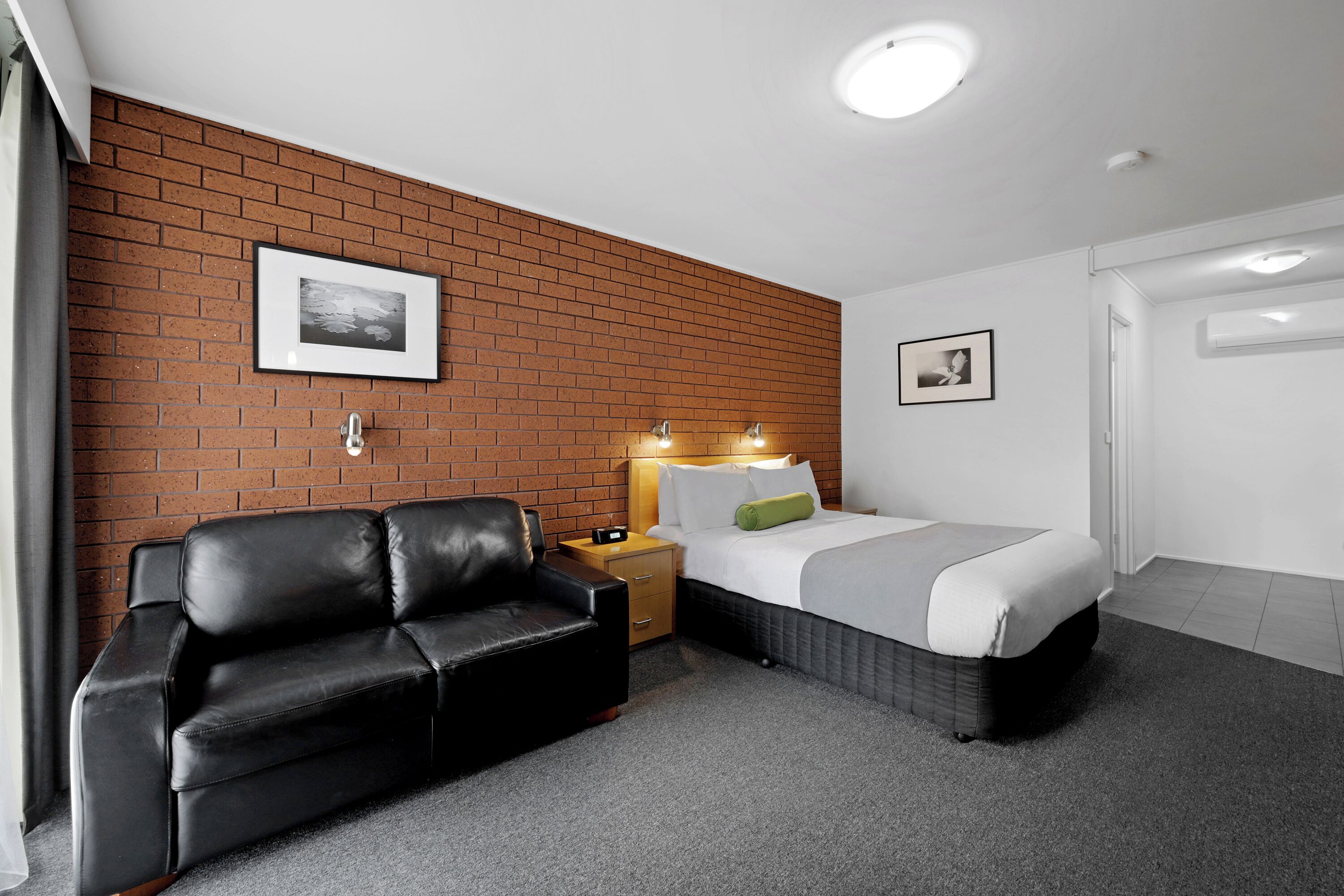 A hotel room with a brick wall, a black leather sofa, a bed with white bedding, and two wall-mounted lights.