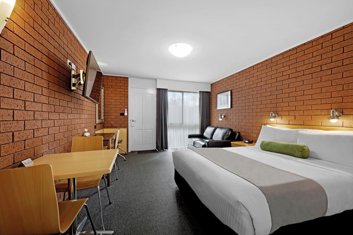 Une chambre d’hôtel avec un mur en briques, un lit avec un oreiller vert, un bureau avec une chaise et une grande fenêtre avec des rideaux.