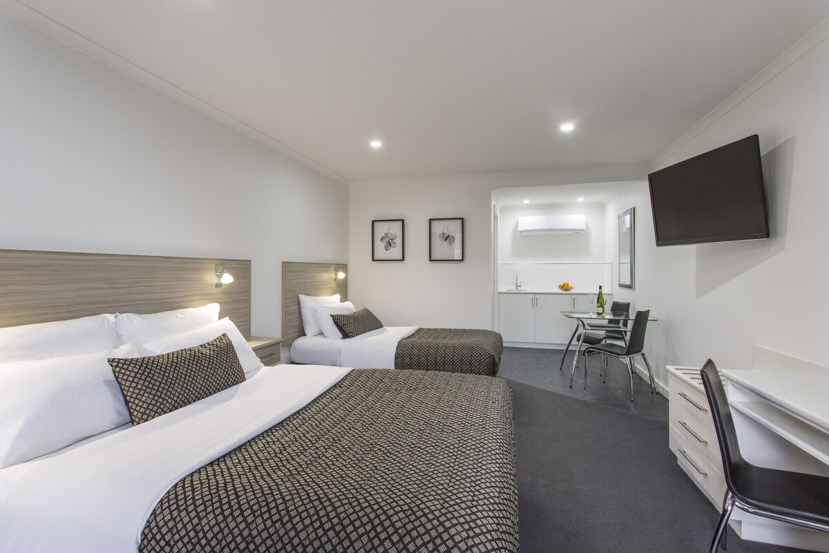 A hotel room with two beds, a desk, a chair, a flat-screen TV, and a kitchenette.