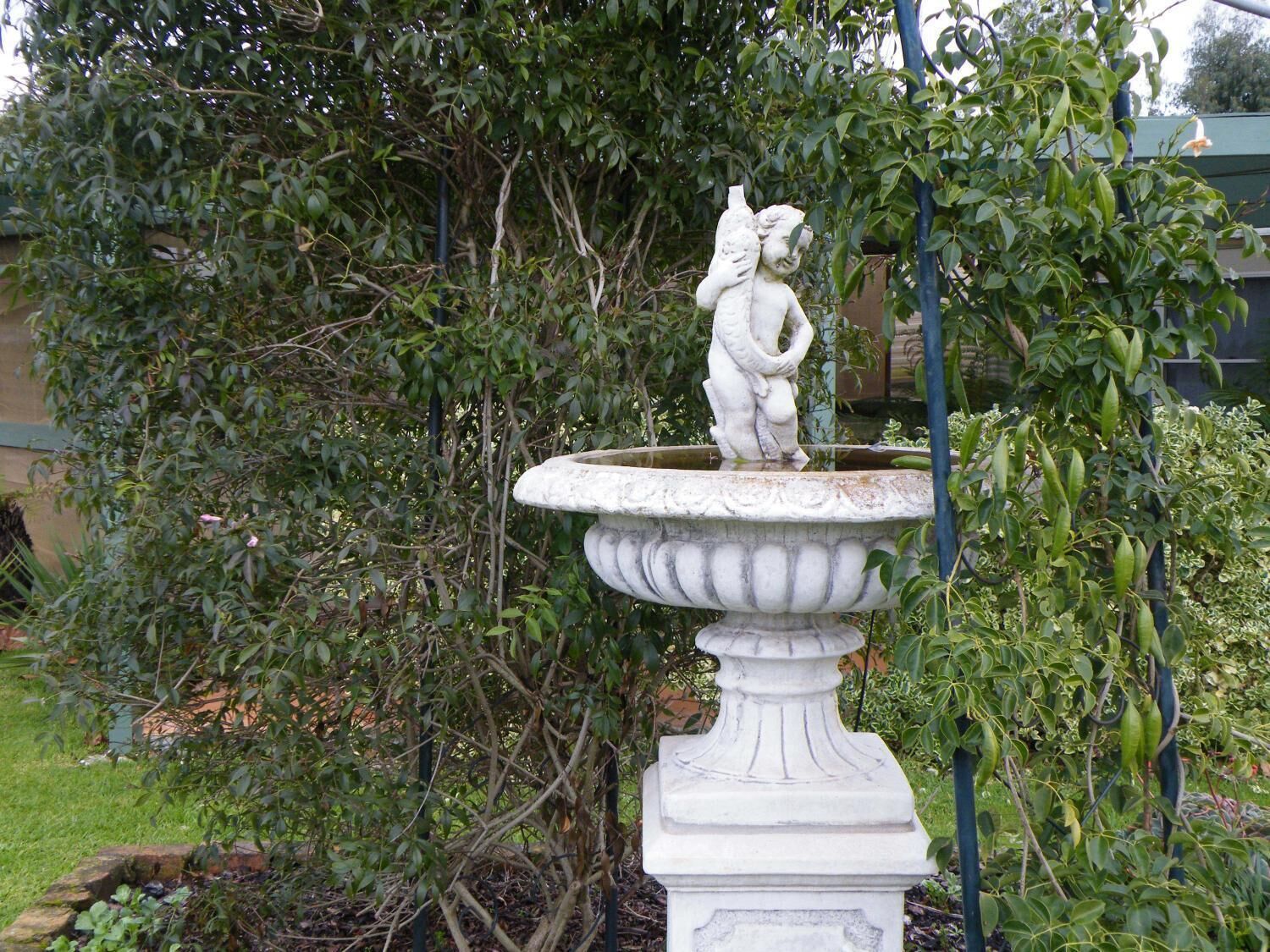 A white stone fountain with a statue of a figure holding a bird, surrounded by lush greenery.