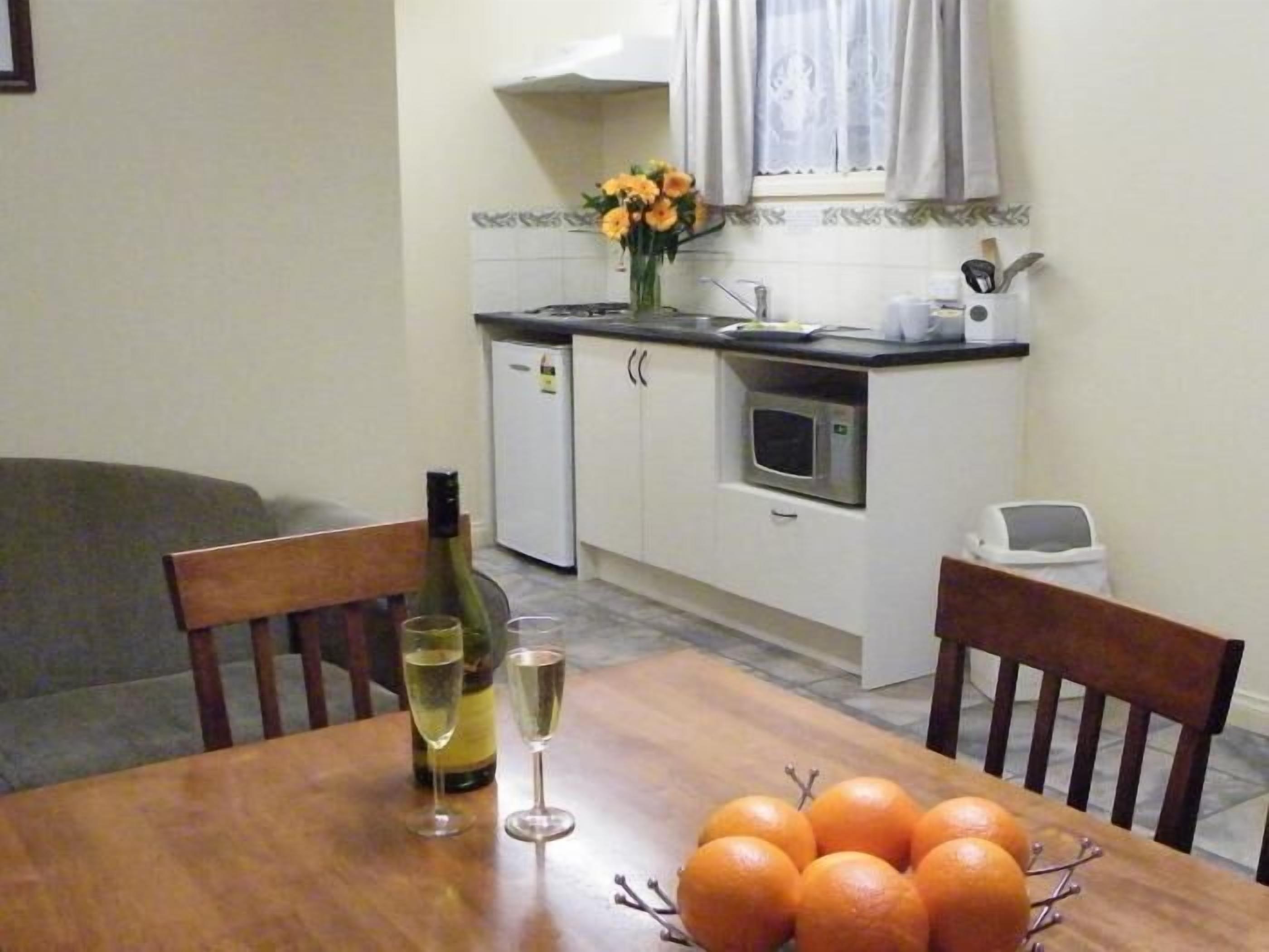 A dining table with two wine glasses and a bottle, a bowl of oranges, and a kitchen in the background.