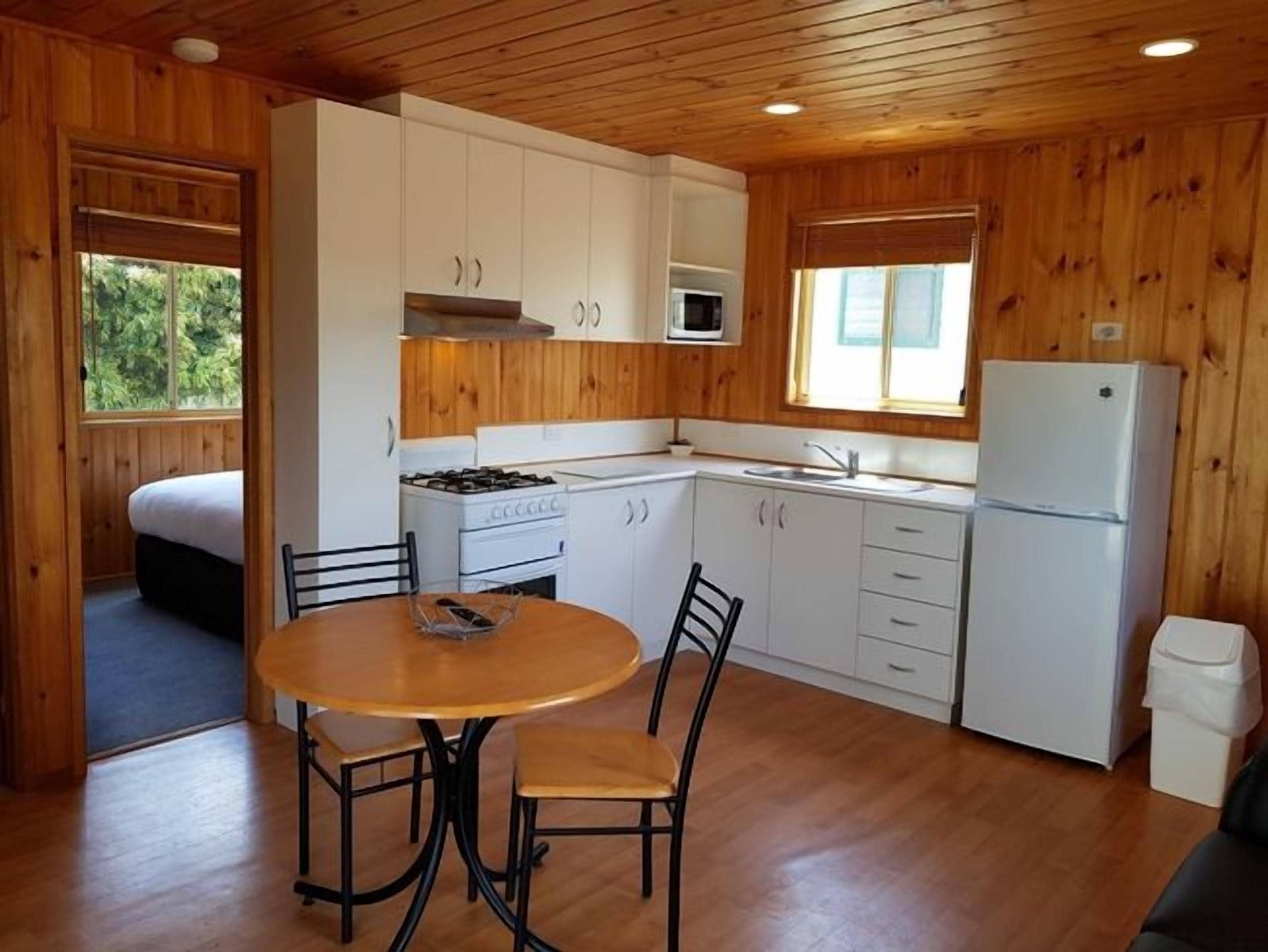 Una cocina compacta con paredes de madera, armarios blancos, una mesa de comedor con sillas y un refrigerador.