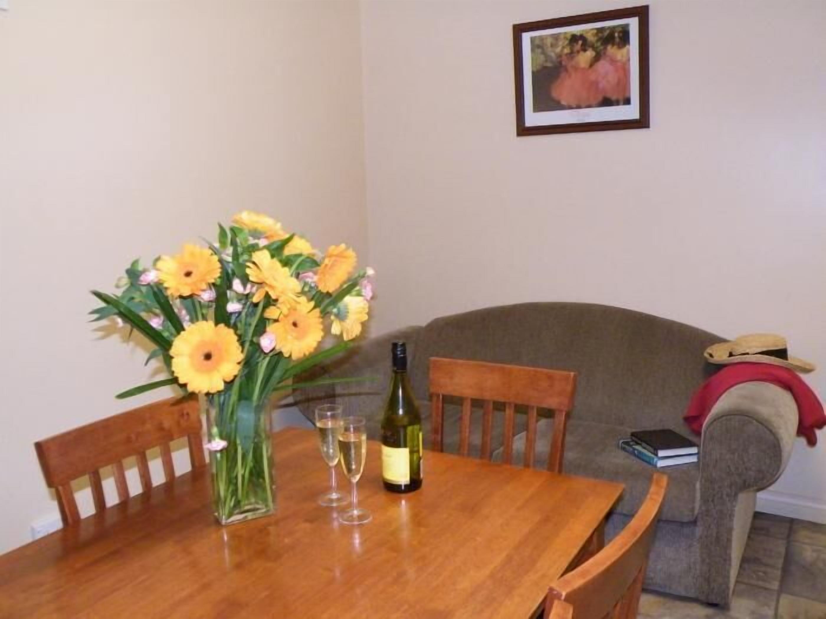 A dining table with a vase of yellow flowers, two wine glasses, and a bottle of wine. There is a framed picture on the wall and a couch in the background.