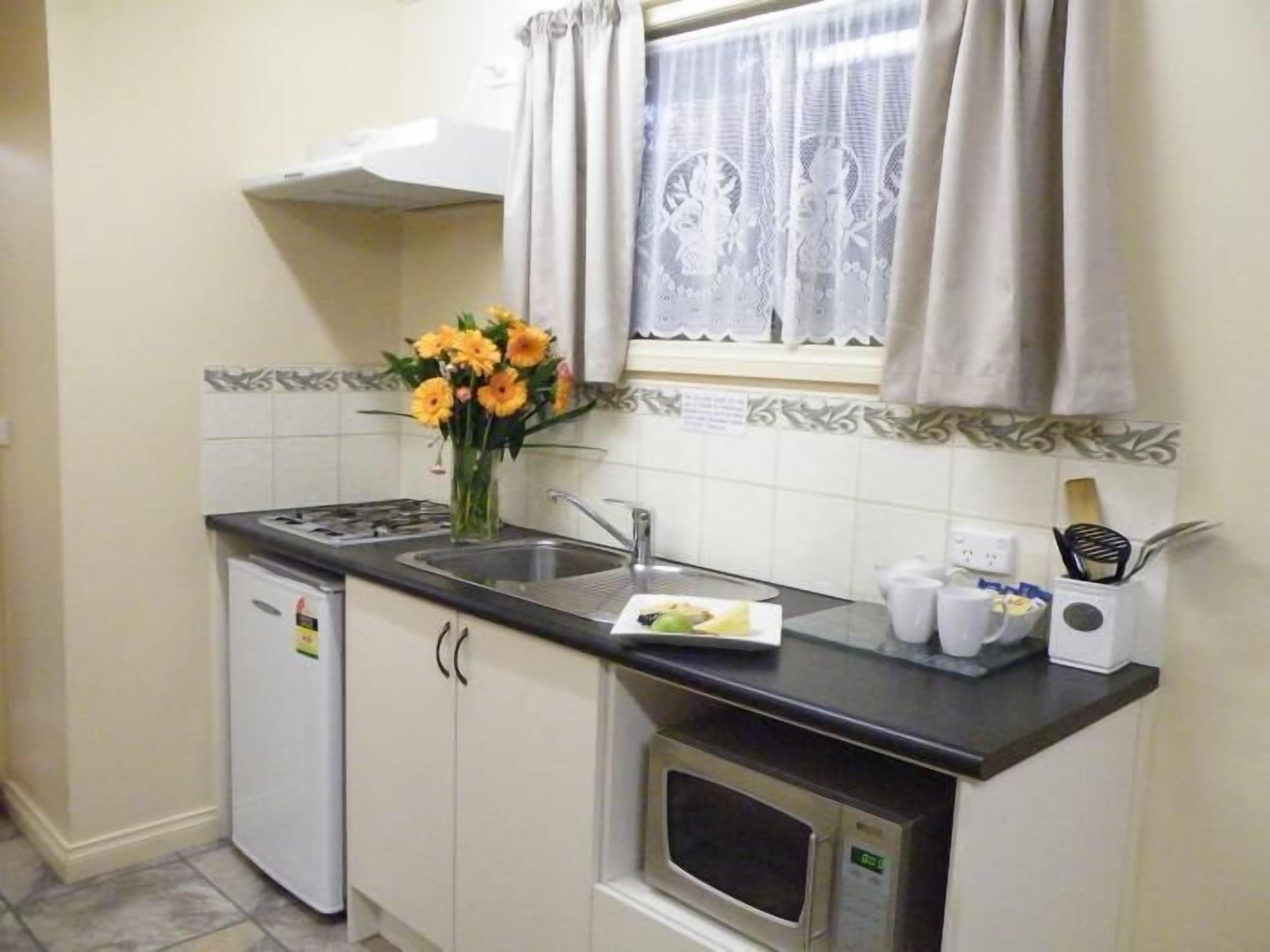 Eine kleine Küche mit Spüle, Herd und Mikrowelle. Auf der Arbeitsfläche steht eine Vase mit Blumen, und es gibt ein Fenster mit Vorhängen.