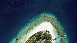 An aerial view of a small island resort with a central building surrounded by lush greenery, a white sandy beach, and clear turquoise waters.