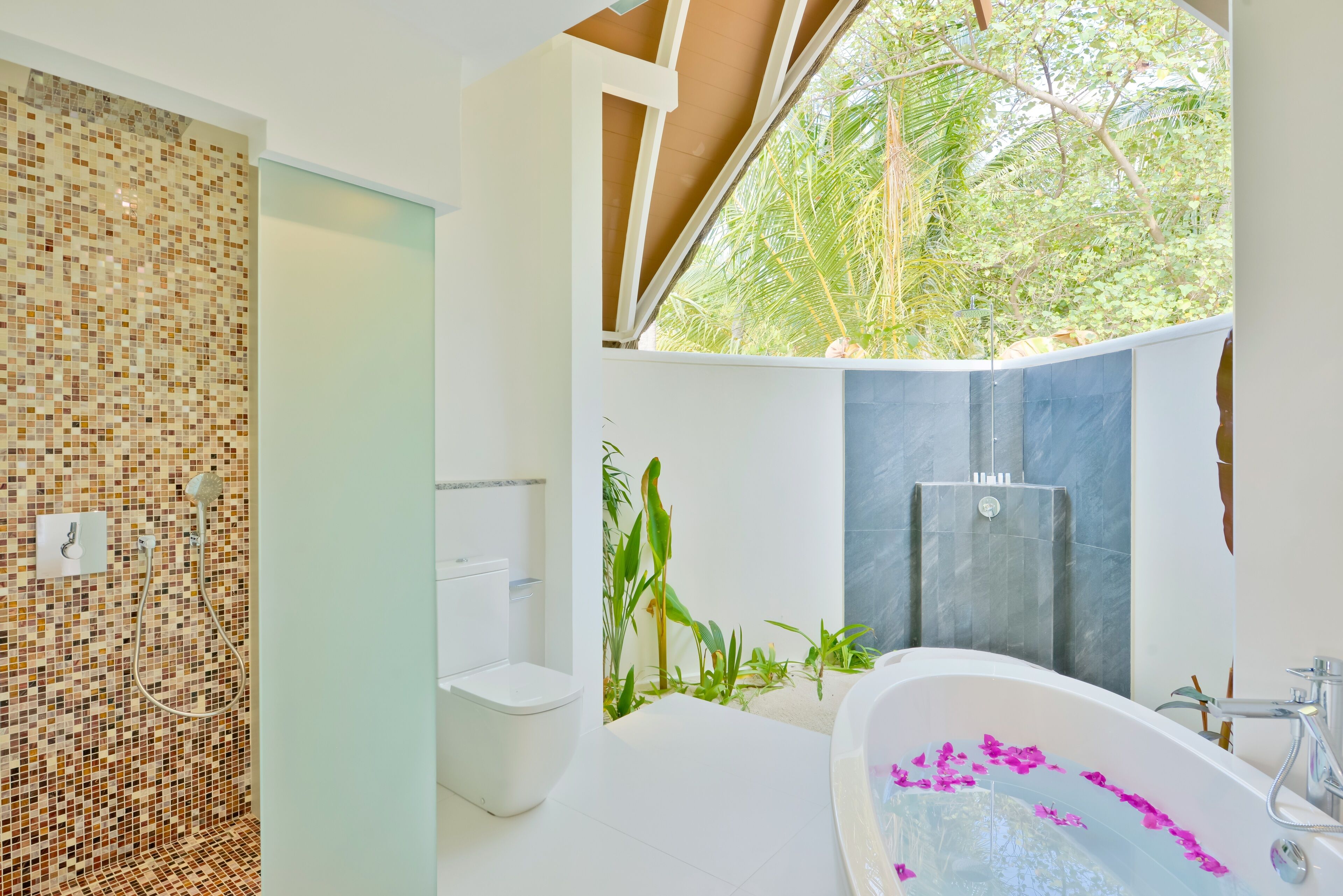 Un bagno moderno con una parete in mosaico, una vasca da bagno indipendente con petali di fiori rosa e uno spazio doccia con vista sul verde.