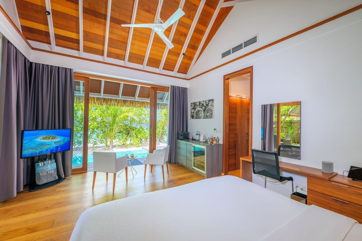 A modern hotel room with a large bed, a desk, a chair, a television, and a view of a pool and greenery.