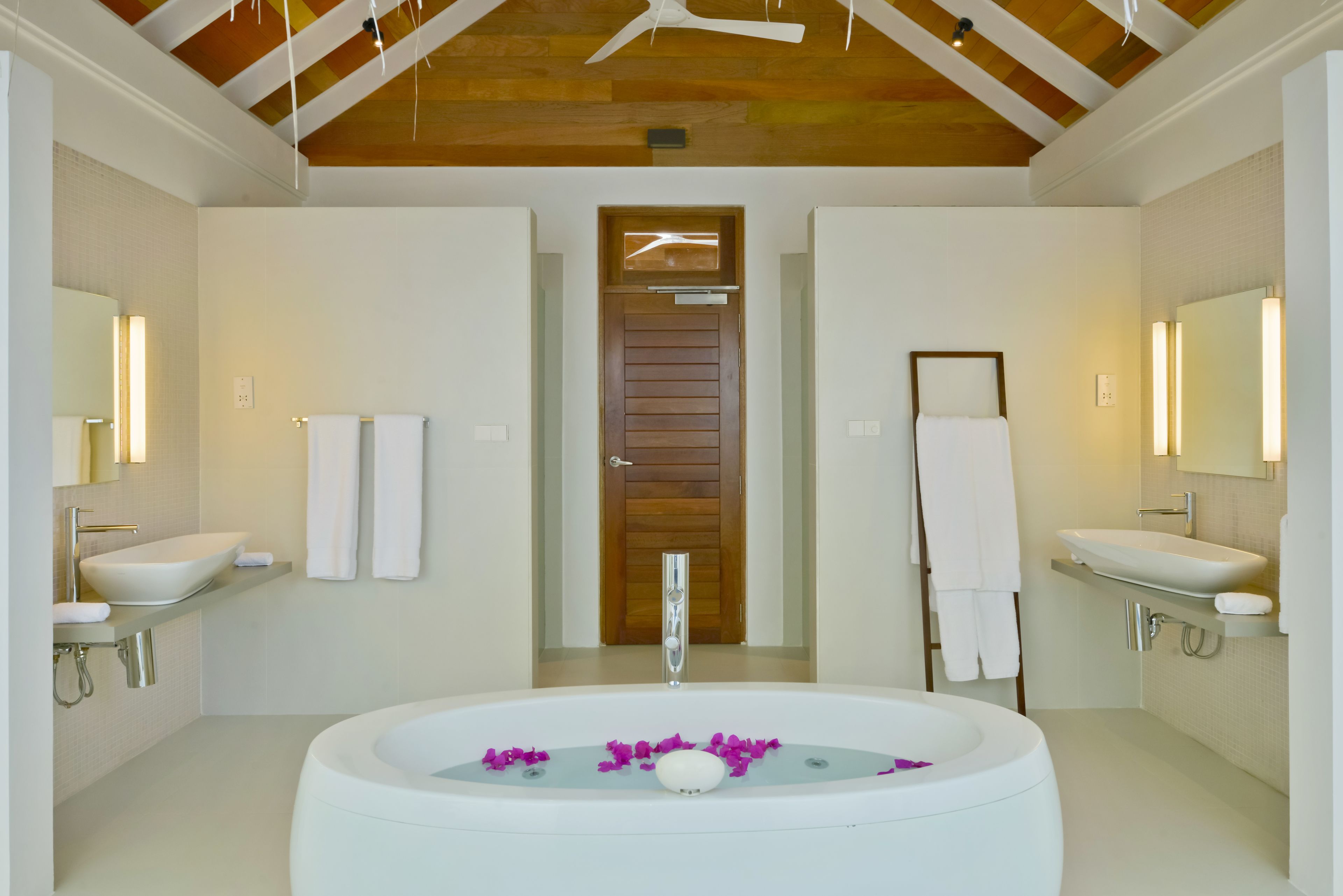A modern bathroom with two sinks, a large bathtub with rose petals, and wooden ceiling beams.