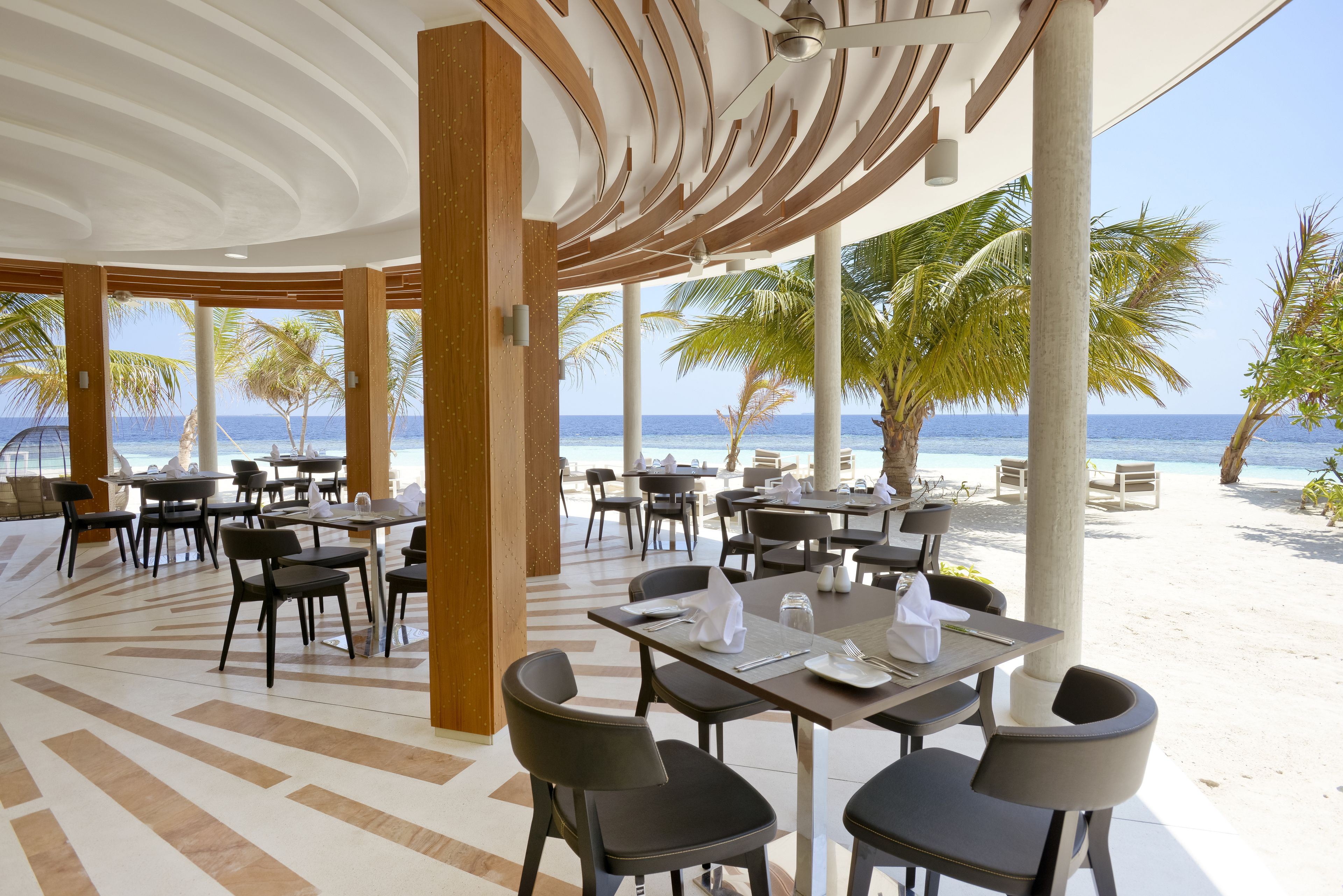Un restaurant situé en bord de mer, avec des tables et des chaises, des palmiers et une vue sur l’océan.
