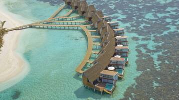 Un resort sobre el agua con bungalows de techo de paja conectados por pasarelas, aguas turquesas cristalinas y una playa de arena.