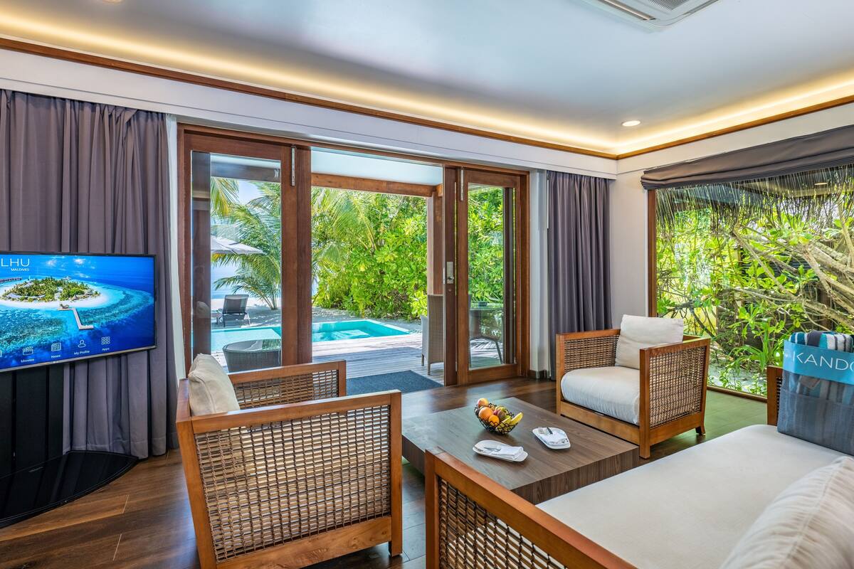 A modern hotel room with a TV, wicker furniture, a dining table, and a view of a pool and greenery.
