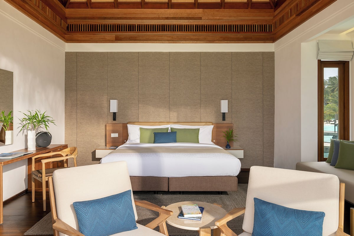A hotel room with a large bed, two bedside tables, a desk, two chairs, and a view of the sea.