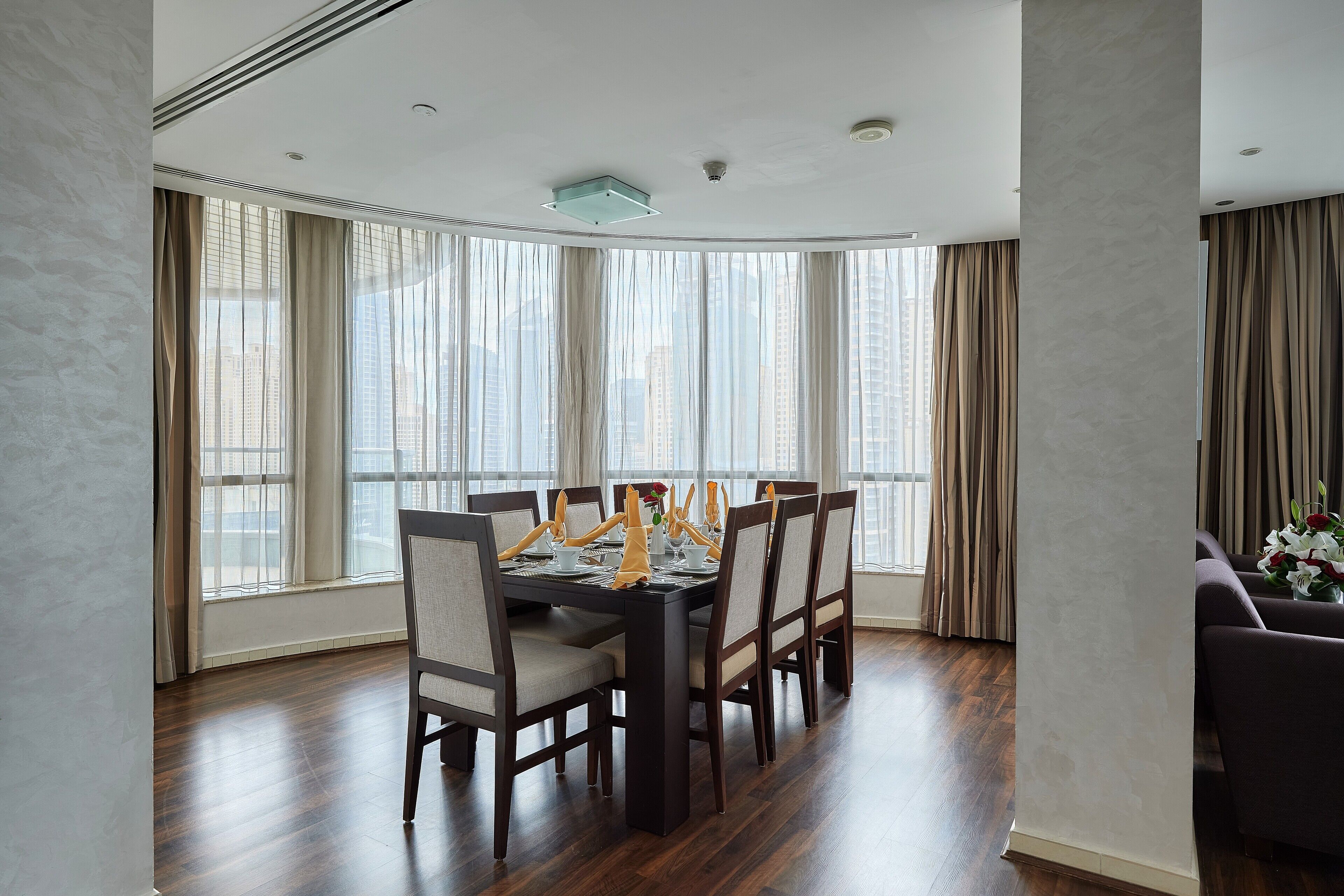 A dining area with a dark wooden table and chairs, large windows with curtains, and a view of tall buildings.
