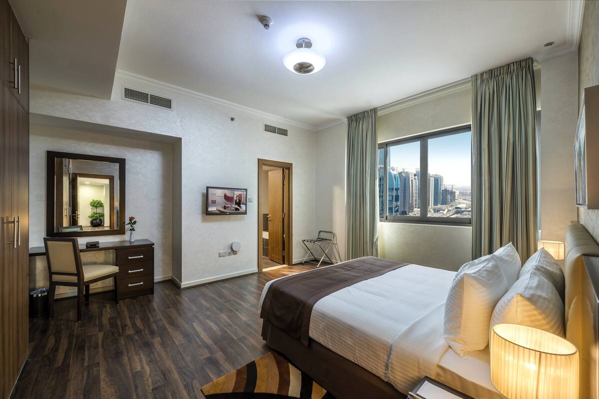 A hotel room with a large bed, a desk with a chair, a mirror, and a view of the cityscape through a large window.