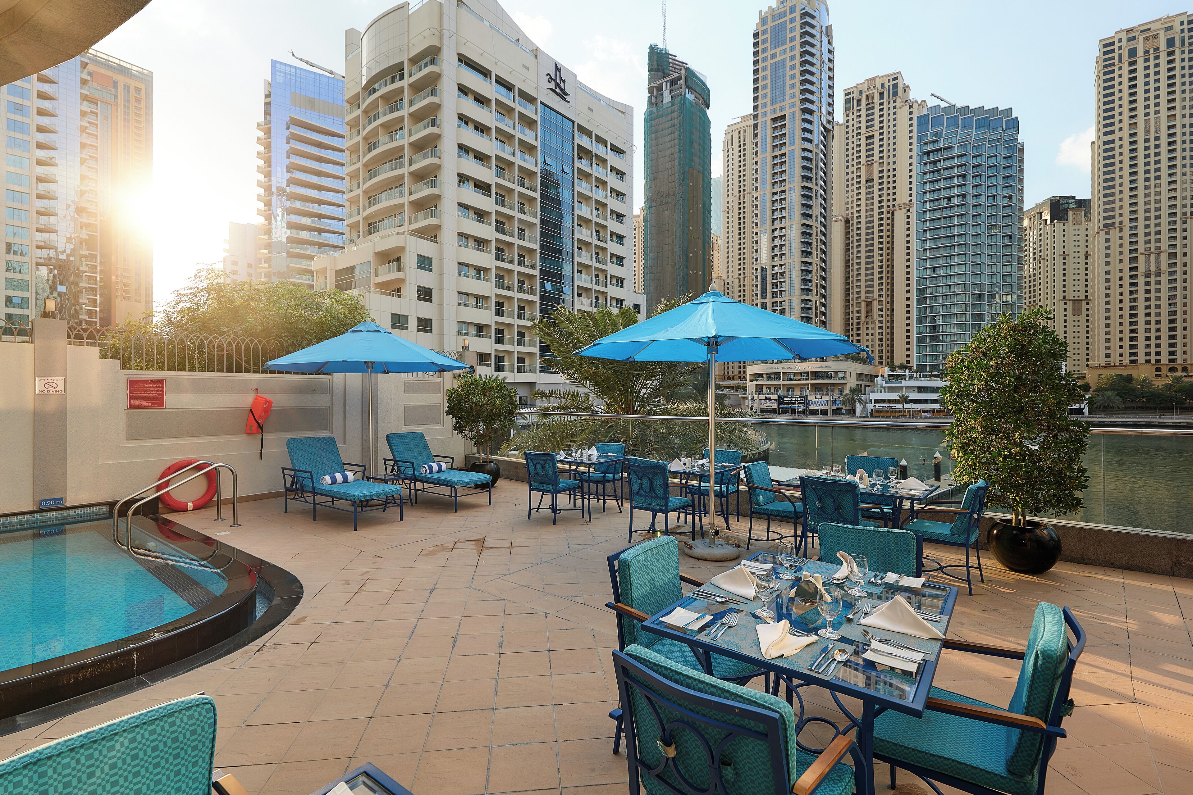 An outdoor pool area with lounge chairs and umbrellas, surrounded by high-rise buildings and a body of water.
