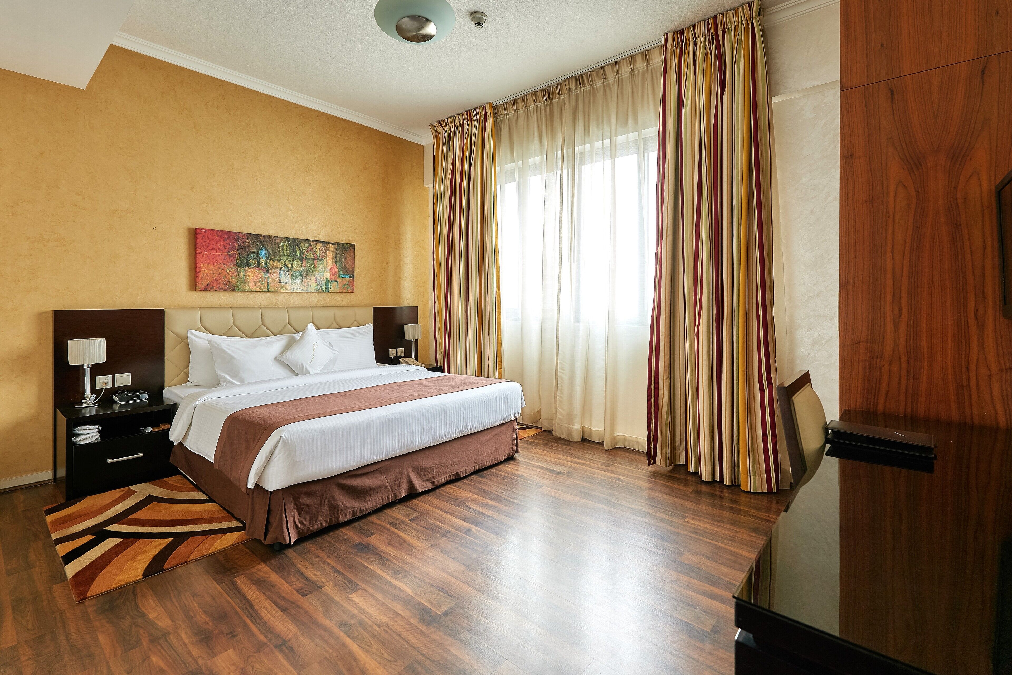 A hotel room with a large bed, a nightstand, a lamp, a painting on the wall, and a window with curtains.