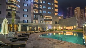 Un área de piscina de hotel con sillas de descanso y sombrillas, una piscina y un edificio moderno al fondo.