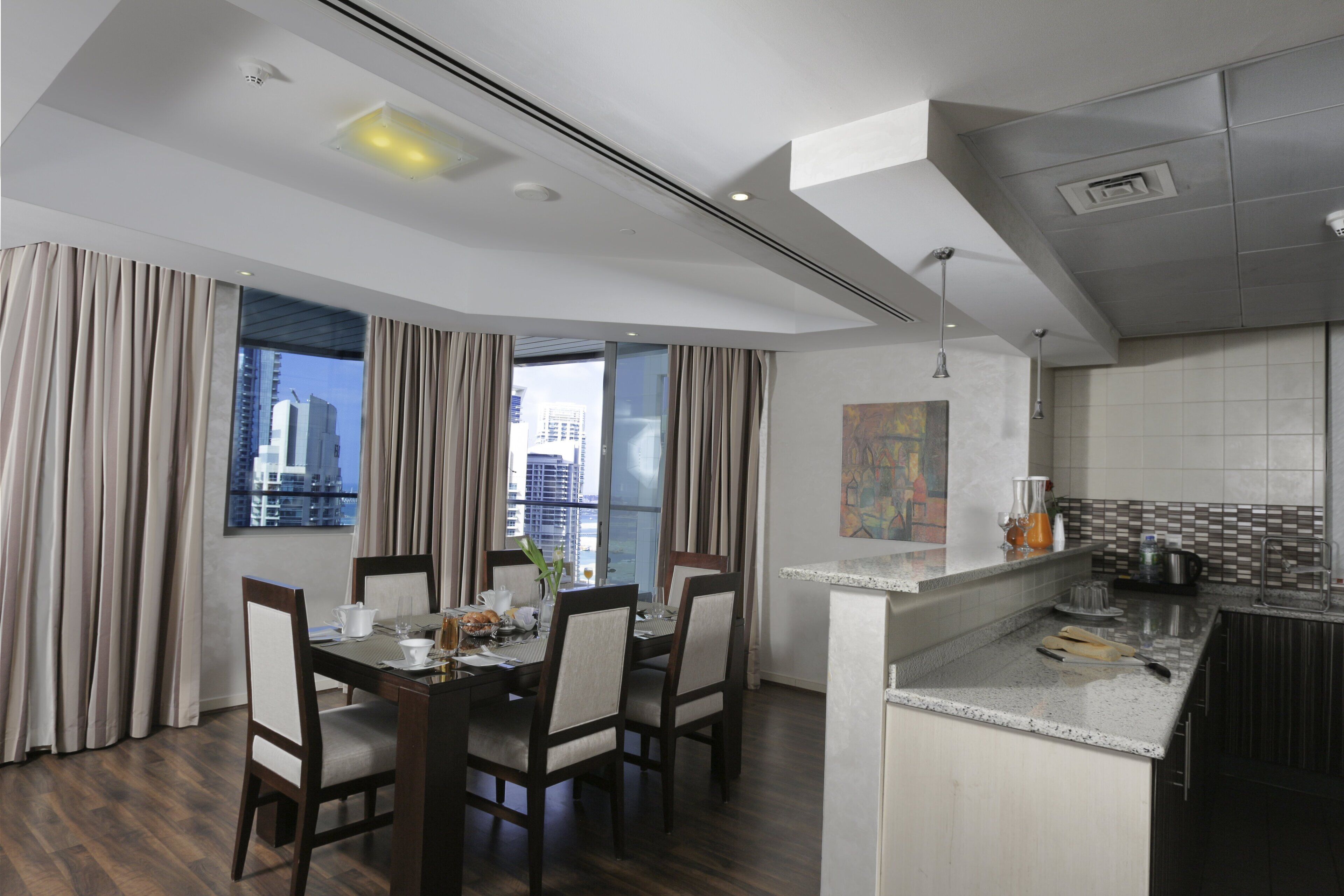 A modern hotel room with a dining area, kitchen, and a view of the cityscape.