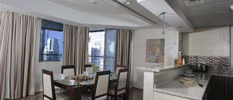 A modern hotel room with a dining area, kitchen, and a view of the cityscape.