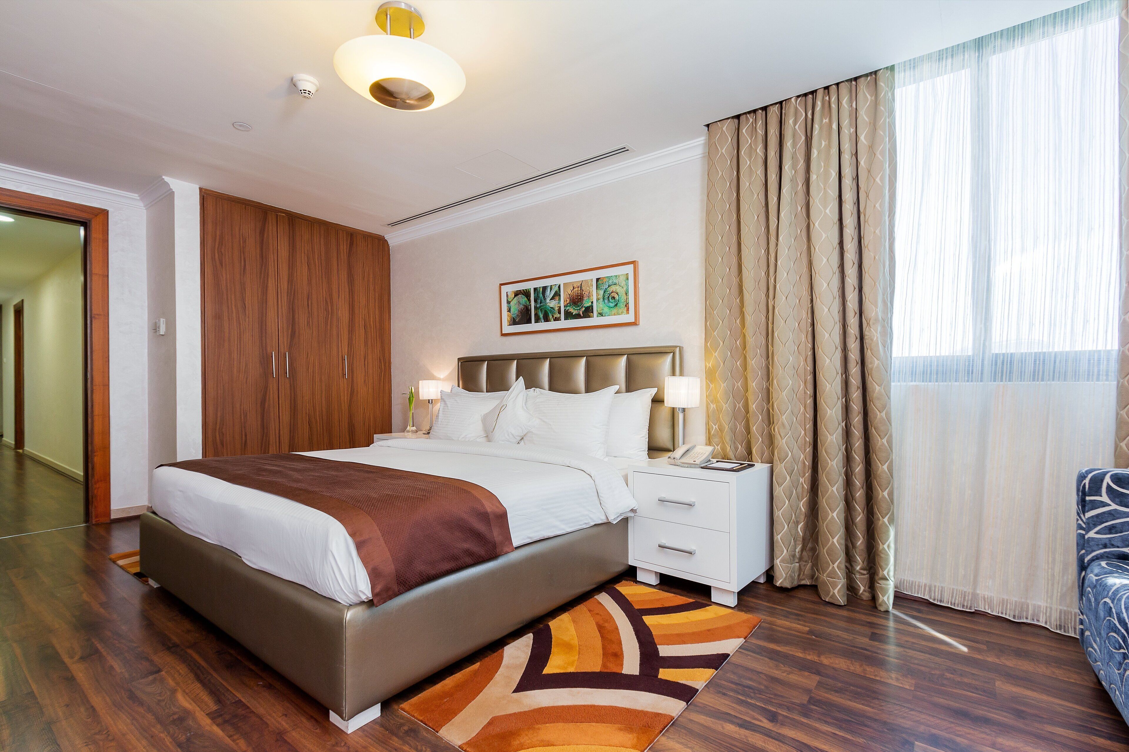 A modern hotel room with a large bed, a bedside table with a lamp, a wardrobe, and a large window with curtains.