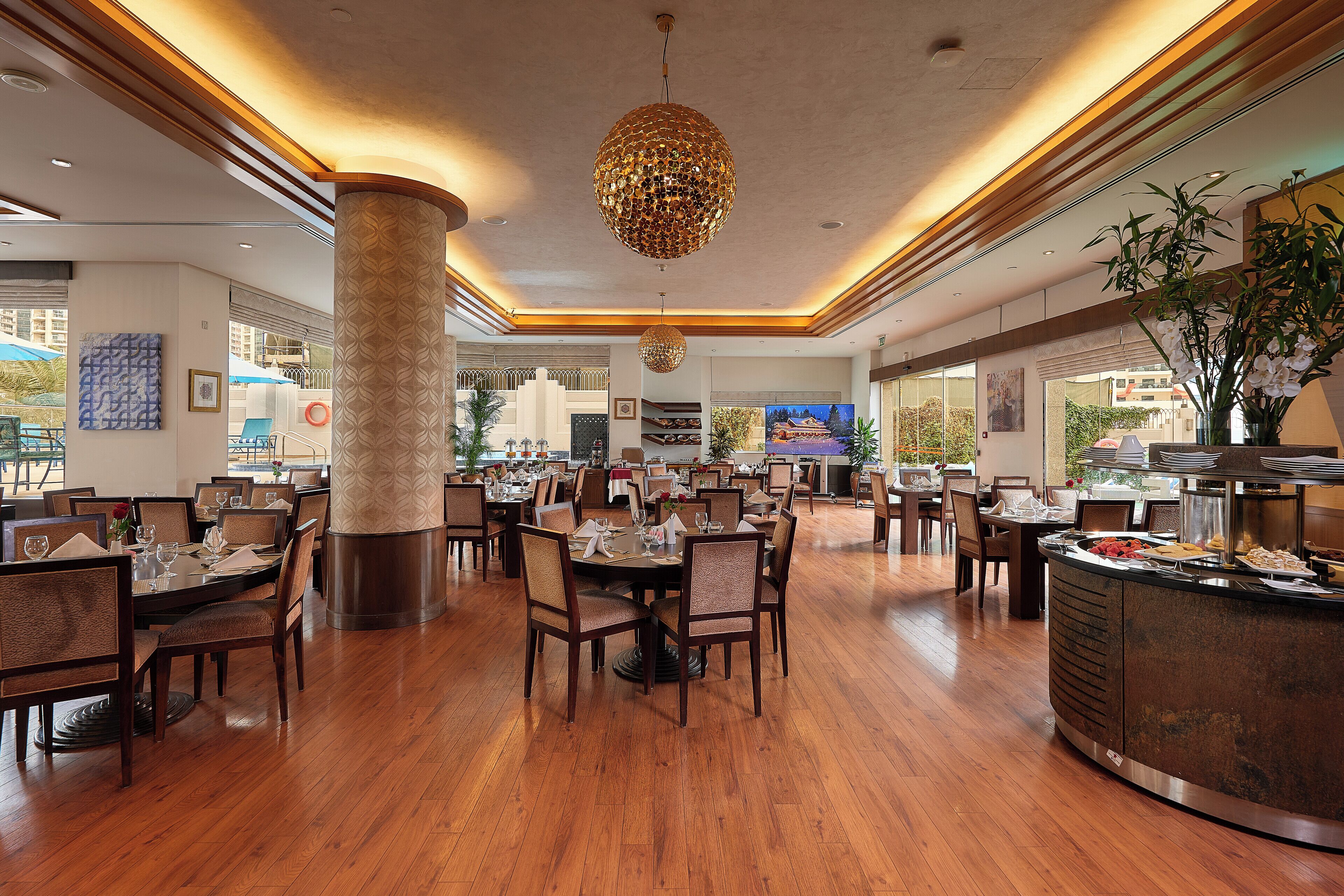 A spacious dining area with wooden flooring, round tables, and chairs. There is a large pillar and a decorative hanging light fixture.