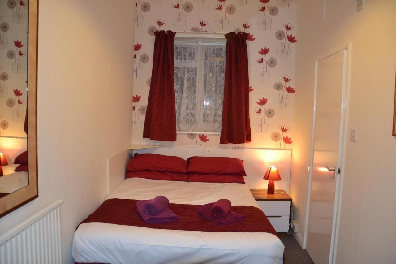 A hotel room with a bed, red curtains, a window with a floral pattern, and two bedside lamps.