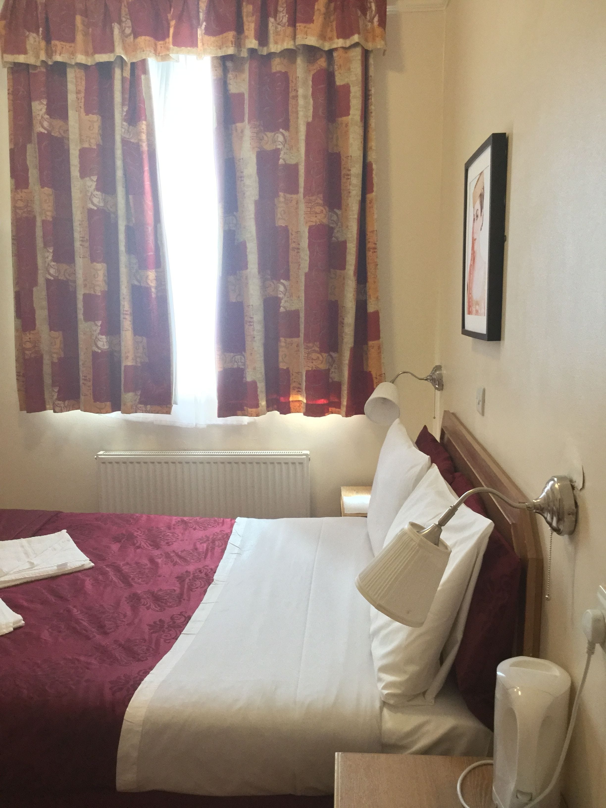 Quarto de hotel com uma cama, luminária, radiador e um quadro na parede.