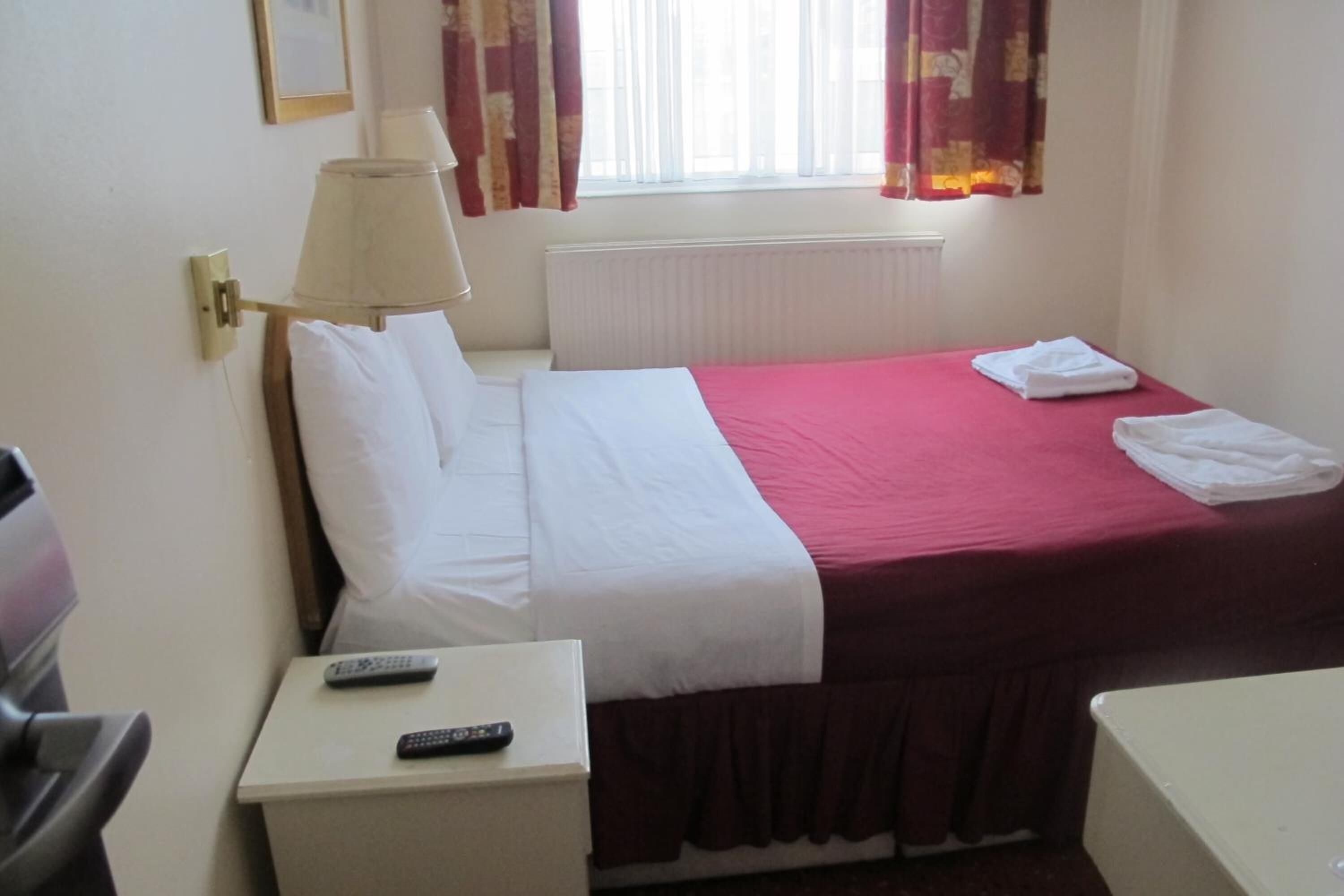 Hotelkamer met een enkel bed, nachtkastje met afstandsbediening, lamp en raam met gordijnen.