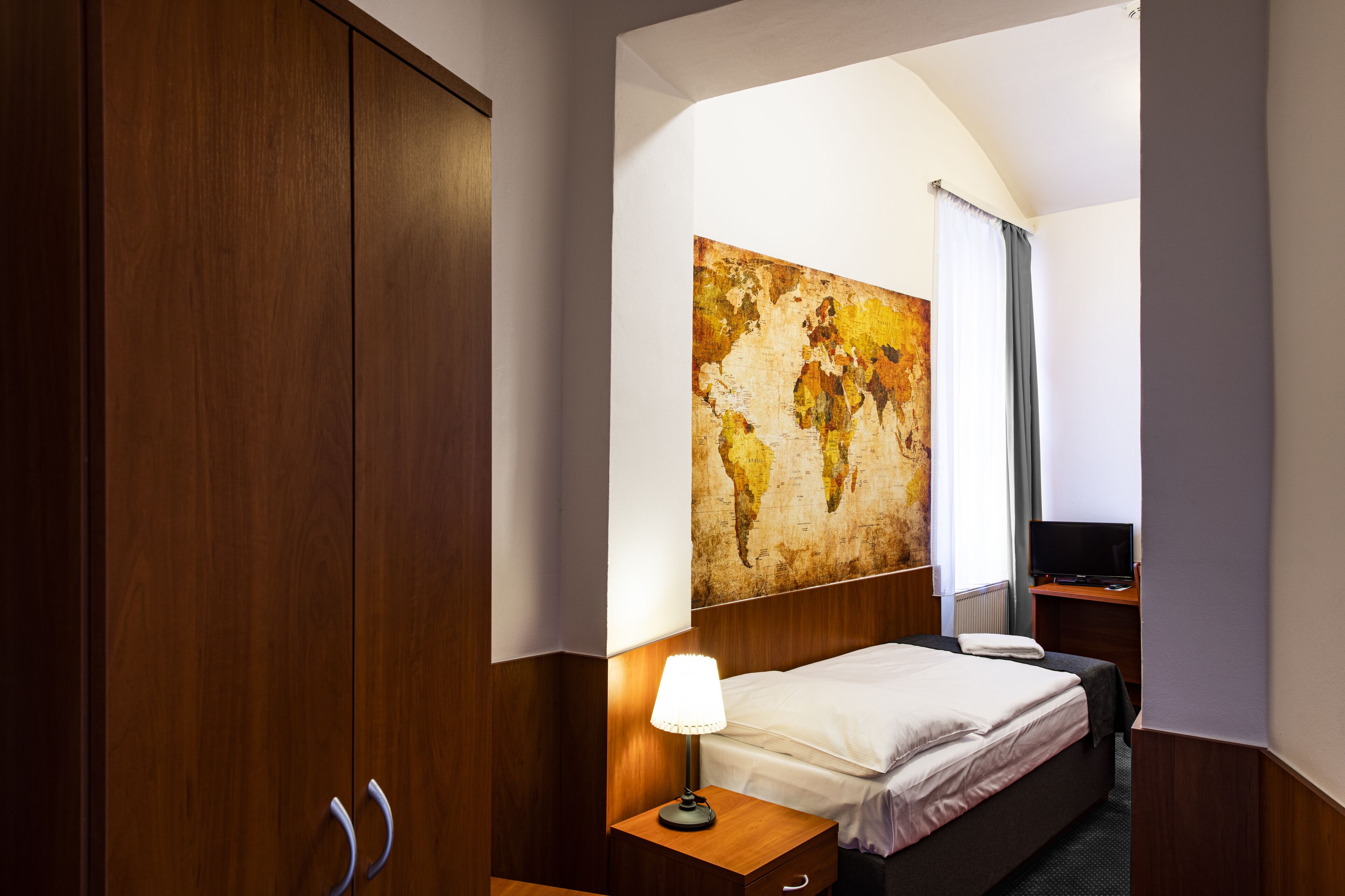 A hotel room with a large wooden wardrobe, a bed with white linens, a nightstand with a lamp, a flat-screen TV, and a world map artwork on the wall.