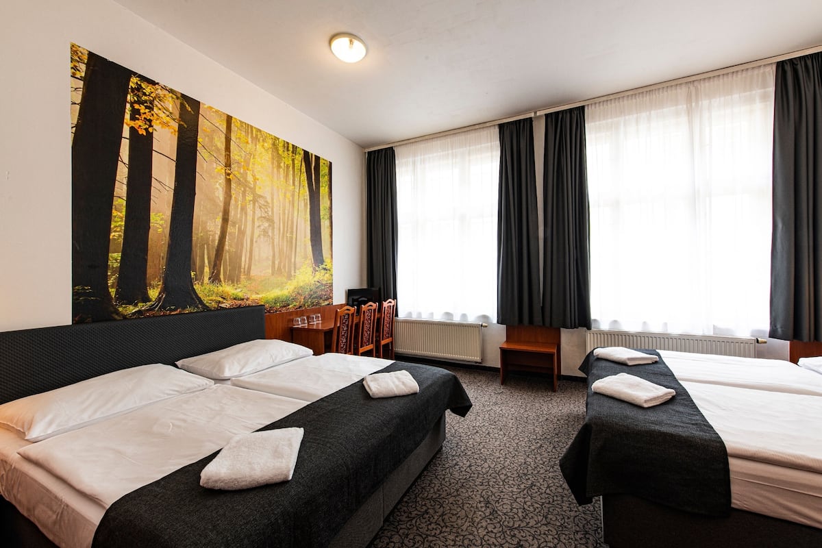 A hotel room with two beds, a large forest-themed painting on the wall, a desk with a chair, and a window with curtains.