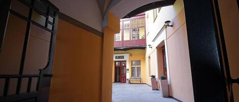 A narrow corridor with a reception desk, a bench, and a door leading to a building with a red door and a balcony.
