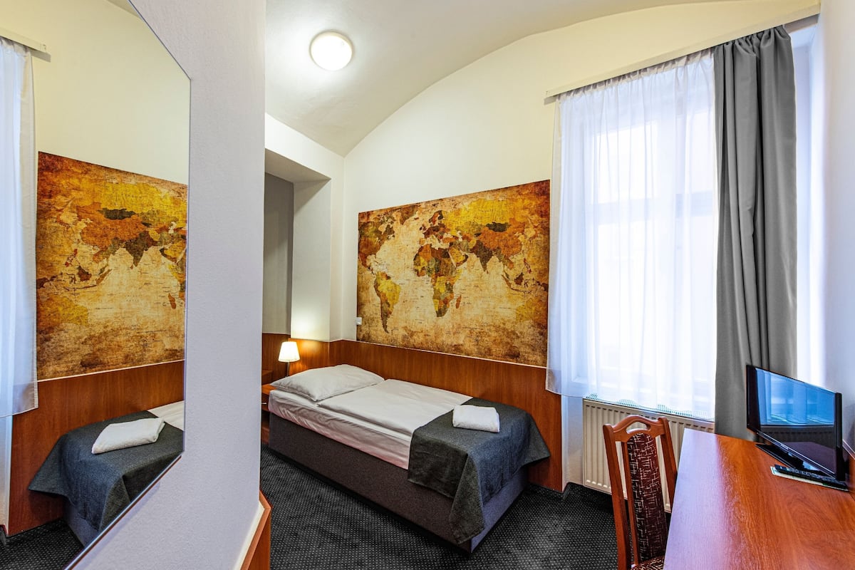 A hotel room with a single bed, a desk with a computer, and two large world maps on the walls.