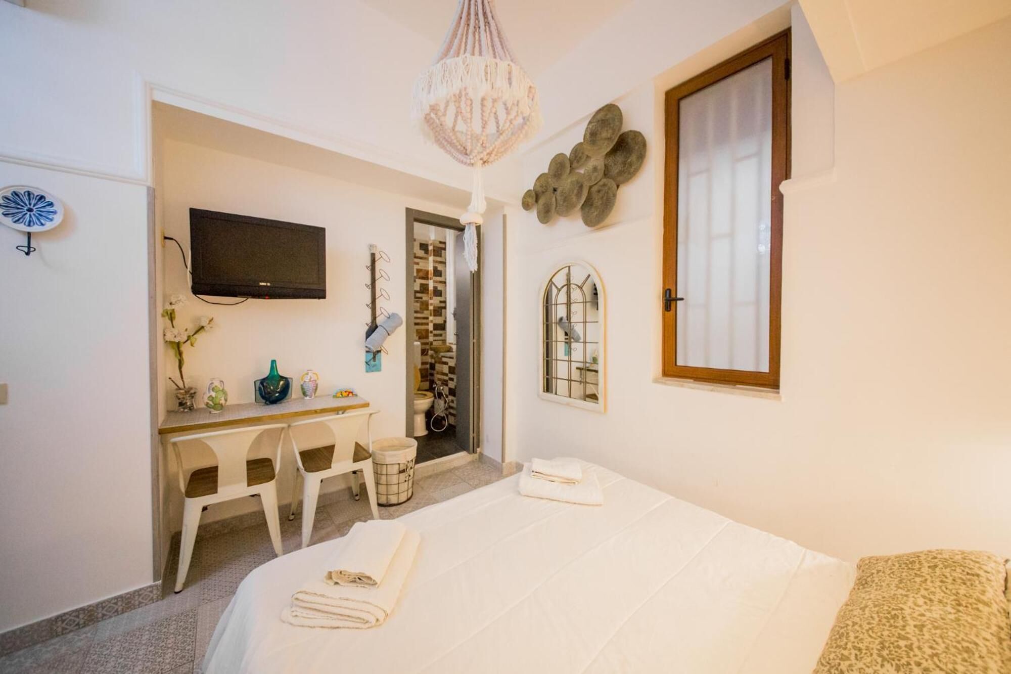 Un dormitorio con una cama, un escritorio y una silla. Hay un televisor en la pared, una ventana y una puerta que da a otra habitación.