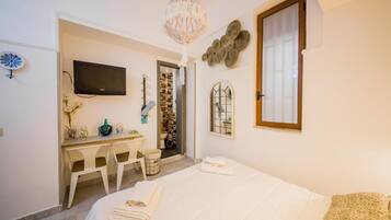 Un dormitorio con una cama, un escritorio y una silla. Hay un televisor en la pared, una ventana y una puerta que da a otra habitación.