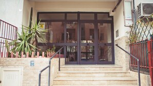 A building entrance with a set of stairs leading up to a glass door, a potted plant, and a red fence. - Casale D'Or (Palermo)