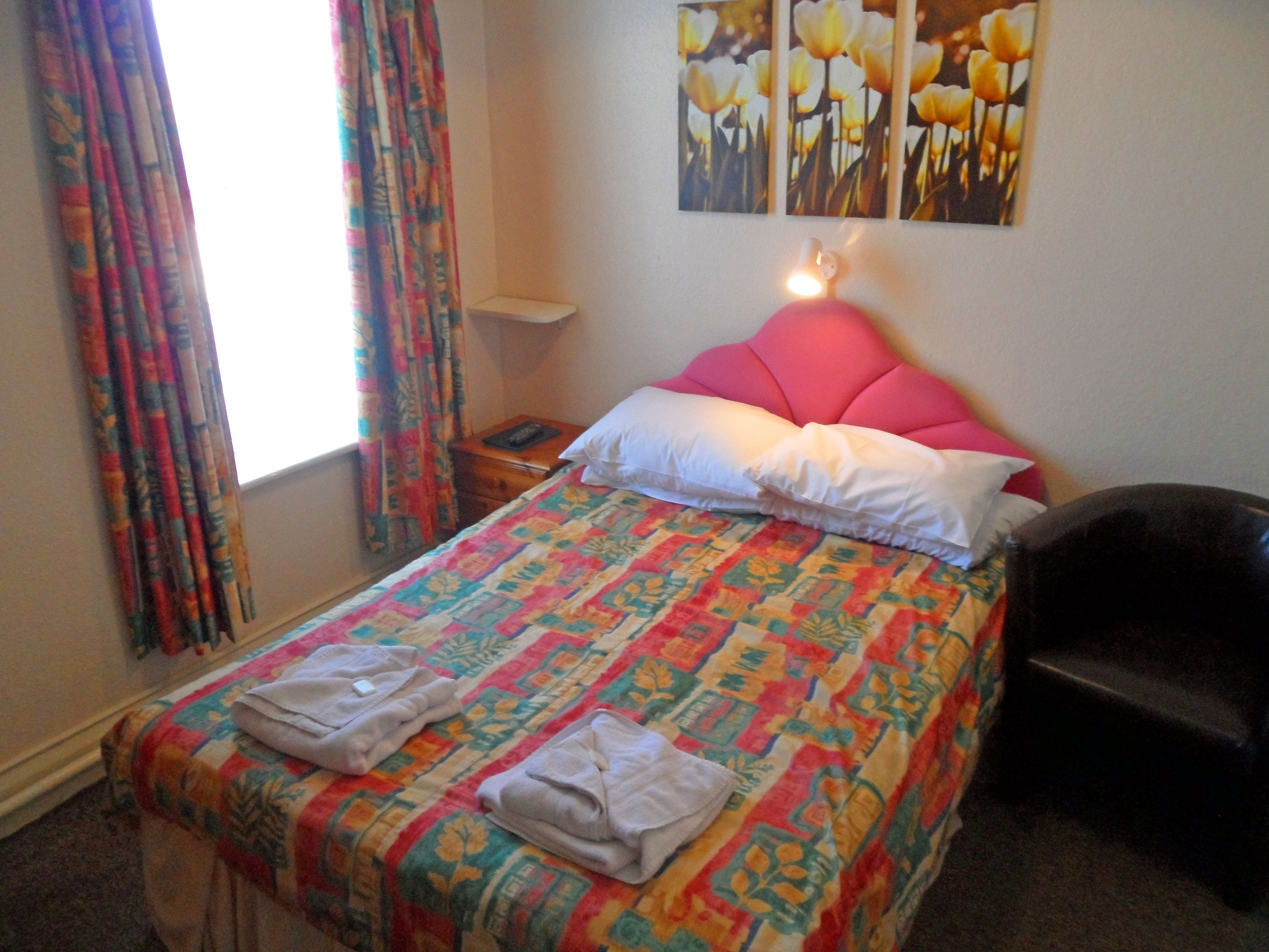 Un letto rifatto con cura, con una coperta colorata, una testata rosa e due cuscini. Si nota un comodino con una lampada, e una sedia nell'angolo.