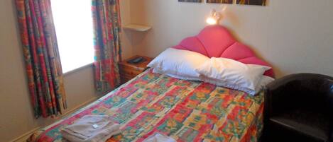 Un letto rifatto con cura, con una coperta colorata, una testata rosa e due cuscini. Si nota un comodino con una lampada, e una sedia nell'angolo.