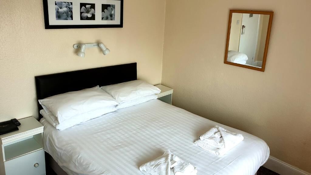 A neatly made bed with white linens and towels, a bedside lamp, a framed picture above the bed, and a mirror on the wall.