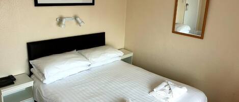 A neatly made bed with white linens and towels, a bedside lamp, a framed picture above the bed, and a mirror on the wall.