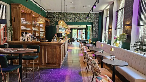 Een bar met houten planken, barstoelen en een toonbank vol flessen en glazen. Er zijn tafels en banken om op te zitten, en de vloer heeft een patroon van tegels.
