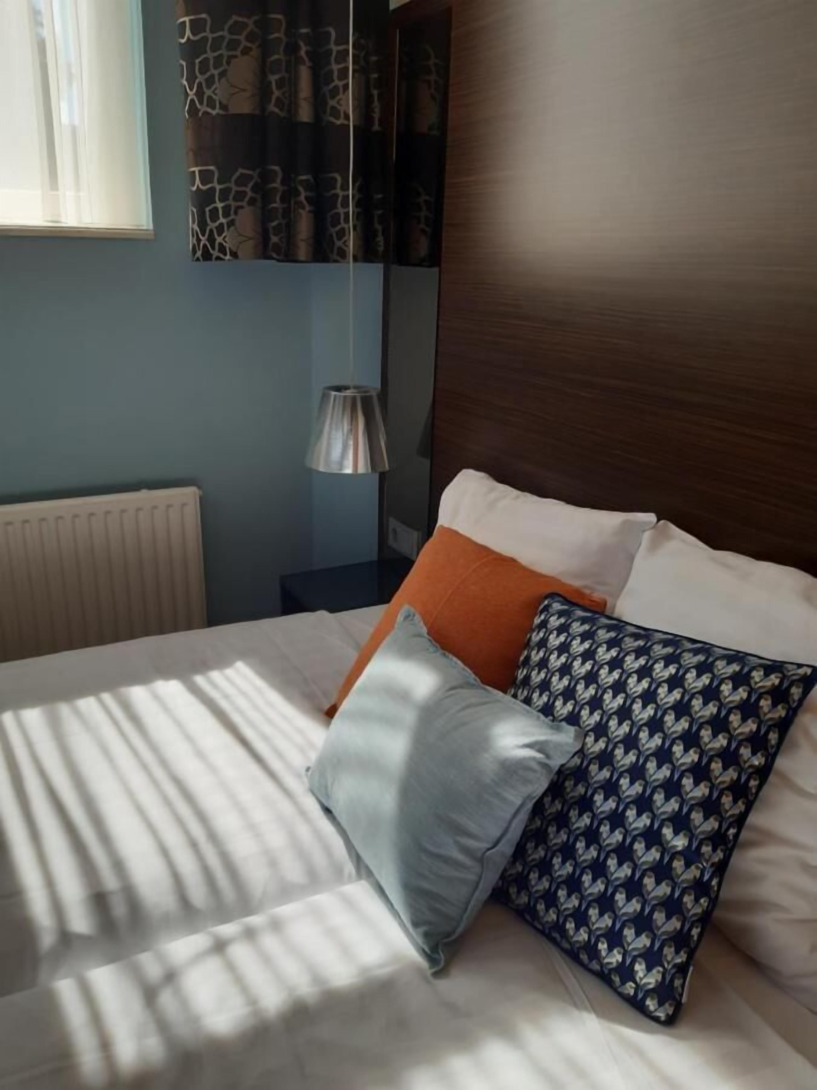 A neatly made bed with white linens and two pillows, one orange and one patterned. A radiator is visible on the wall, and a hanging lamp is above the bed.