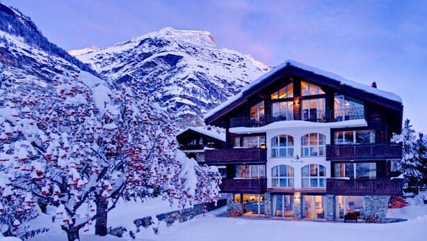 Un hôtel à plusieurs étages, avec une montagne enneigée en toile de fond, de grandes fenêtres et une façade en pierre.