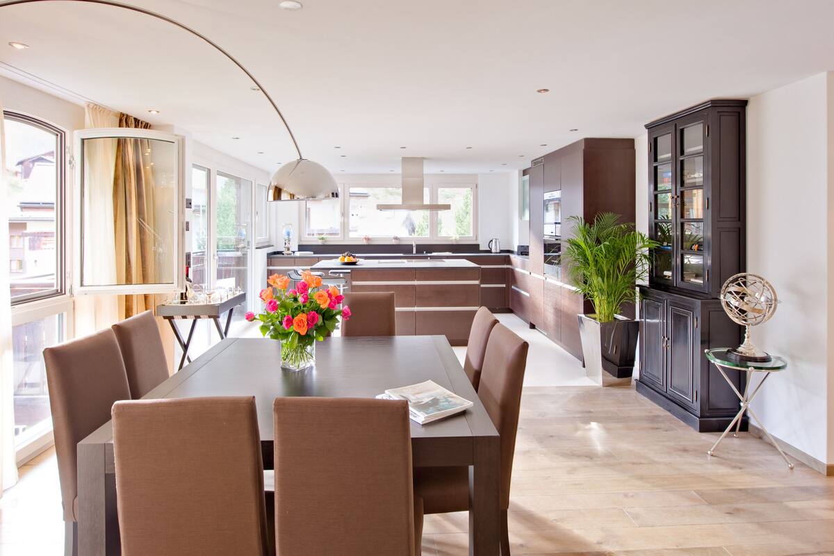 A modern kitchen with a dining area, featuring a central island, a built-in oven, and a large window providing natural light.