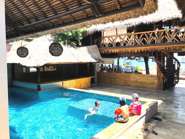 Una piscina con techo de paja, asientos de madera y vista al mar.
