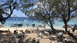 A beachside café with outdoor seating, tables, and chairs, overlooking the sea with boats in the background.