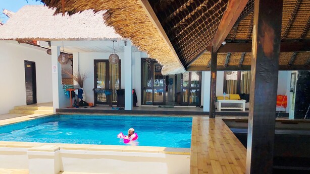 A swimming pool with a person in a pink float, surrounded by a covered area with wooden flooring and a thatched roof.